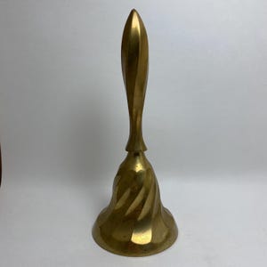 May include: A gold-toned brass bell with a twisted, pointed handle. The bell has a wide, flared base and a swirling pattern.