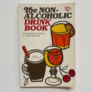 May include: The cover of a vintage cookbook titled "The Non-Alcoholic Drink Book" by Dolores M. Du Nah & H. Jean Groudle. The cover features illustrations of three non-alcoholic drinks: a mug of hot chocolate, a glass of orange juice, and a glass of a red drink with a slice of orange.