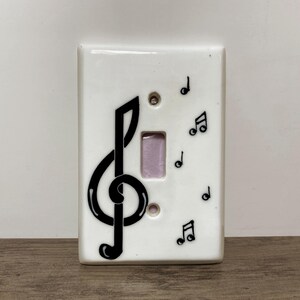 May include: A rectangular white light switch plate featuring a large black treble clef symbol on the left side and several smaller black musical notes scattered across the right side. The plate has a central rectangular cutout for a toggle switch.