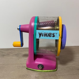 May include: A colorful pencil sharpener with a clear plastic chamber, featuring a pink base, a blue and yellow handle, and a purple dial. The text "yikes!" is printed on a blue sticker on the side.
