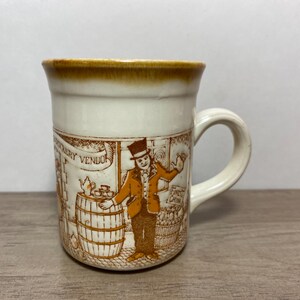 May include: A white ceramic mug with a brown rim and handle. The mug features a vintage illustration of a street scene with a man selling fruit and another man selling tea. The text "FINEST CHINA" and "GROCERY VENDOR" are visible on the mug.