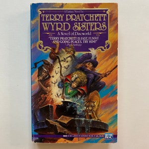 May include: A book cover with a purple background and a colorful illustration of a group of people and a cat. The title of the book is "Wyrd Sisters" by Terry Pratchett. The text on the cover reads "A Novel of Discworld" and "Terry Pratchett is fast, funny and going places. Try him!" - Piers Anthony.
