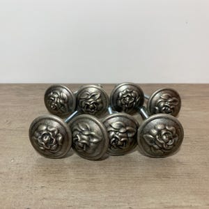 Peut inclure: Un ensemble de six boutons de placard ronds en métal avec une finition argent vieilli. Chaque bouton présente un motif floral en relief représentant une rose sur la face avant et comprend une tige filetée métallique à l'arrière.
