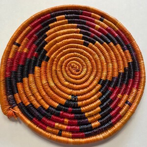 Peut inclure: Un dessous de plat rond en osier avec un motif en spirale dans des tons d'orange, de rouge et de noir.