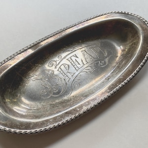 May include: Oval silver bread tray with ornate detailing. The word "BREAD" is engraved in elegant script across the centre. The tray has a beaded edge and a slightly tarnished finish, giving it a vintage appearance.