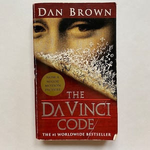 May include: A red paperback book with the title "The Da Vinci Code" by Dan Brown. The cover features a close-up of a man's eye and a piece of parchment with writing on it. The book is a #1 worldwide bestseller.