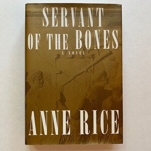 Peut inclure: Un livre à couverture rigide intitulé "Servant of the Bones: A Novel" par Anne Rice. La couverture est de couleur sépia avec le titre et le nom de l'auteur en blanc, en gras. L'arrière-plan présente une image estompée de personnages.
