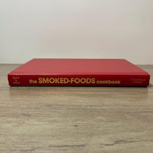 May include: A red cookbook titled "the SMOKED-FOODS cookbook" with gold lettering. The book is closed and lying flat on a wooden surface. The publisher's name, Park & Park, is printed on the spine.