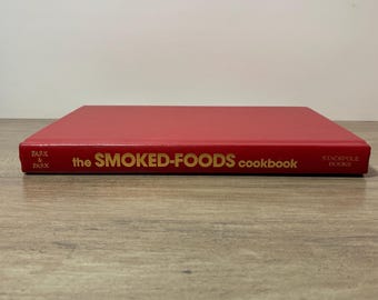 May include: A red cookbook with the title "the SMOKED-FOODS cookbook" in gold lettering. The book is closed and resting on a wooden surface. The publisher's name, Park & Park, is printed on the spine.