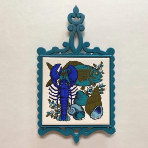 Peut inclure: Sous-plat en céramique bleu sarcelle avec un bord festonné et une poignée décorative. Le sous-plat présente une tuile blanche avec un homard bleu, un poisson turquoise et des coquillages marrons.