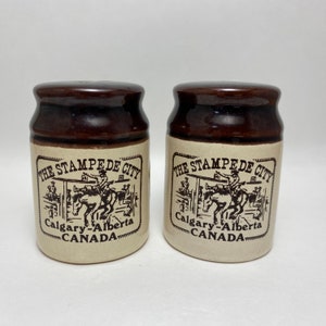 May include: Two brown ceramic salt and pepper shakers with a white label that reads "The Stampede City Calgary-Alberta CANADA". The label features a black and white illustration of a cowboy riding a horse.