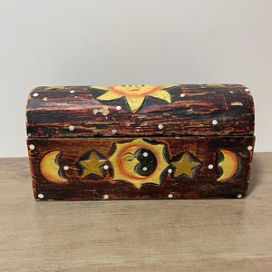 以下が含まれることがあります： 木製の宝箱で、天体のデザインが施されています。 宝箱は、太陽、月、星が黄色とオレンジ色で、濃い茶色の背景に描かれています。 白い点が上部に散らばっています。 宝箱は、湾曲した蓋と長方形のベースを持っています。