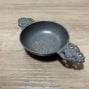 May include: A small, antique pewter tea caddy with ornate handles and a decorative floral pattern in the bowl's center. The caddy is a dark gray color and sits on a wooden surface.