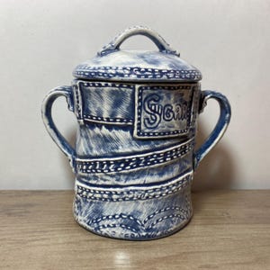 May include: A blue and white ceramic sugar bowl with a lid and two handles. The design resembles denim, with stitched details and the word "Sugar" in a rectangular frame. The bowl is on a wooden surface.