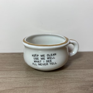 May include: A small, white ceramic cup with a handle and a brown rim. The cup has the text "KEEP ME CLEAN USE ME WELL WHAT I SEE I'LL NEVER TELL" printed on the front. The cup is sitting on a wooden surface.