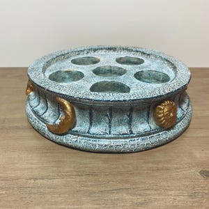 May include: A round, teal-colored holder with a cracked texture and six circular openings on the top surface. The exterior side features raised, gold-colored celestial motifs, including a crescent moon and a sun face, arranged around the perimeter.