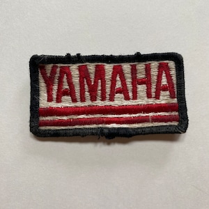 May include: A white and red embroidered patch with the word "YAMAHA" in red lettering. The patch has a black border and two red stripes.