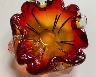 May include: A red and orange glass ashtray with a wavy, flower-like design. The ashtray is made of clear glass with a red and orange ombre effect.