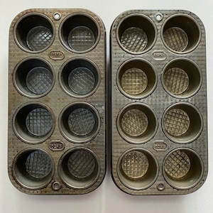 May include: Two vintage metal muffin tins with 12 individual cups. The tins are gray and have a textured surface. Each cup has a raised, patterned bottom. The tins are labeled "EKCO" and "OVERLY" on the sides.