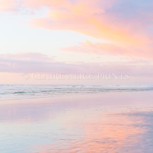 May include: A pastel pink and orange sunset reflected in the calm waters of a beach. The sky is a soft pink and orange, with clouds that are a light blue. The water is a light blue, with a few ripples on the surface. The sand is a light brown.