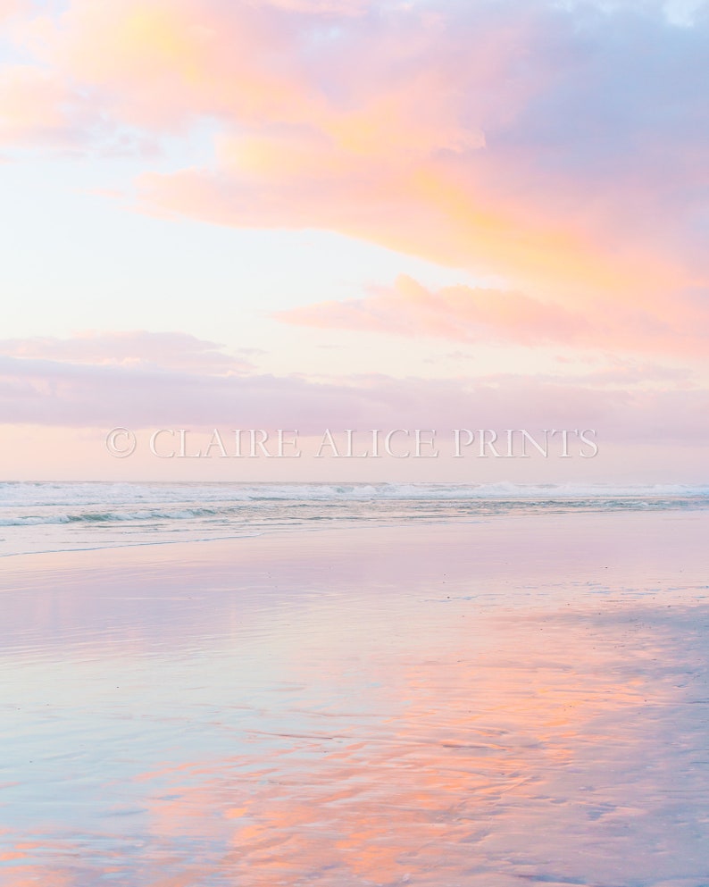 May include: A pastel pink and orange sunset reflected in the calm waters of a beach. The sky is a soft pink and orange, with clouds that are a light blue. The water is a light blue, with a few ripples on the surface. The sand is a light brown.