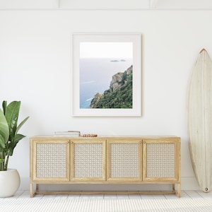 May include: A white framed photograph of a coastal landscape with a blue ocean and green cliffs. The photograph is hanging on a white wall above a light wood cabinet with four doors. A white surfboard is leaning against the wall to the right of the cabinet.