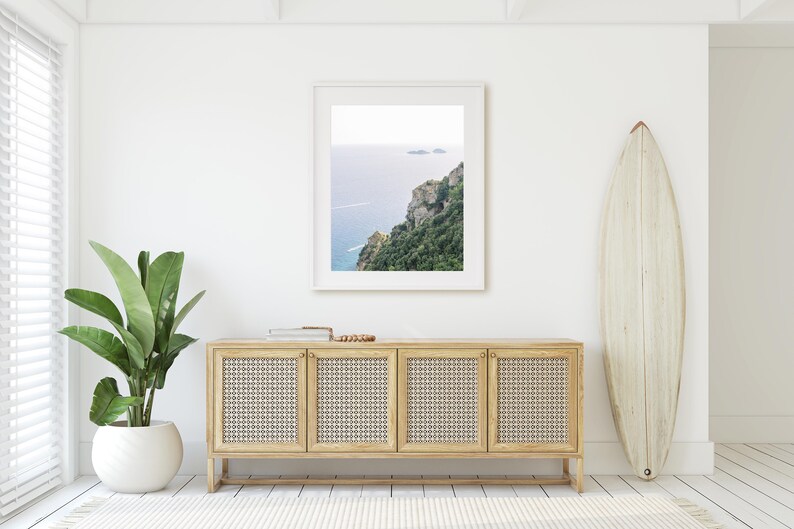 May include: A white framed photograph of a coastal landscape with a blue ocean and green cliffs. The photograph is hanging on a white wall above a light wood cabinet with four doors. A white surfboard is leaning against the wall to the right of the cabinet.