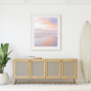 May include: A white framed picture of a pink and purple sunset over a calm ocean with a reflection of the sky in the water. The picture is hanging on a white wall above a light wood cabinet with four doors. A white surfboard is leaning against the wall to the right of the cabinet.