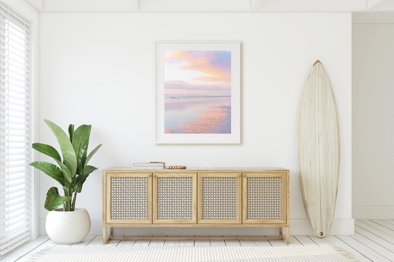 May include: A white framed picture of a pink and purple sunset over a calm ocean with a reflection of the sky in the water. The picture is hanging on a white wall above a light wood cabinet with four doors. A white surfboard is leaning against the wall to the right of the cabinet.