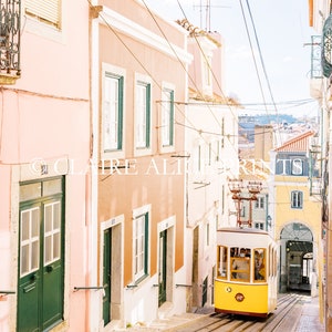 May include: A yellow vintage tram travels up a steep, cobblestone street lined with pastel pink and peach buildings. The tram has a white roof and windows, and the front of the tram is decorated with a yellow circle with a number 1 inside.