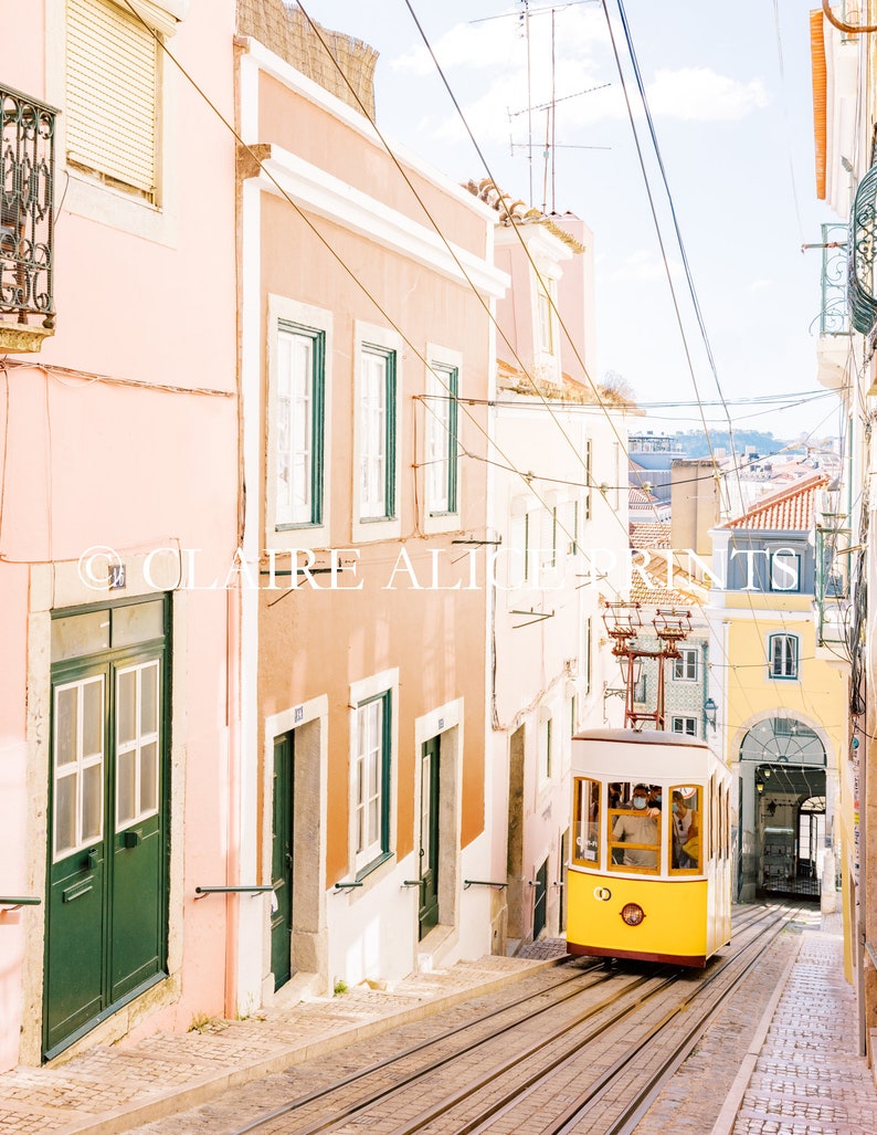 May include: A yellow vintage tram travels up a steep, cobblestone street lined with pastel pink and peach buildings. The tram has a white roof and windows, and the front of the tram is decorated with a yellow circle with a number 1 inside.