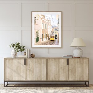 May include: A framed photograph of a yellow tram traveling up a narrow street in a city. The buildings on either side of the street are painted in shades of pink and yellow.