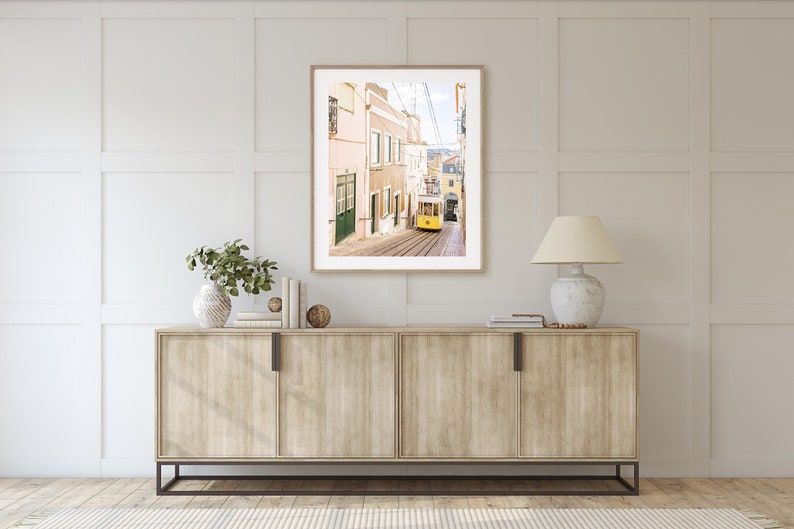 May include: A framed photograph of a yellow tram traveling up a narrow street in a city. The buildings on either side of the street are painted in shades of pink and yellow.