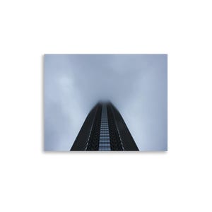 May include: A tall, dark skyscraper with a grid-like facade of windows tapering toward the top, disappearing into a thick, overcast gray sky. The building is captured from a low-angle perspective, emphasizing its height and vertical lines.