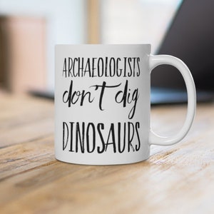 May include: White ceramic coffee mug with black text that reads "Archaeologists don't dig dinosaurs".