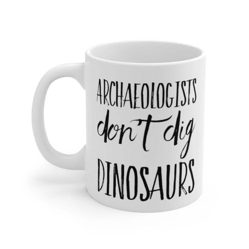 May include: White ceramic coffee mug with black text that reads "Archaeologists don't dig dinosaurs".