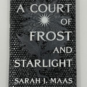 May include: A book cover for "A Court of Frost and Starlight" by Sarah J. Maas. The cover is black and encrusted with small, sparkling black gems. The title and author's name are in white, with a detailed gray floral design.