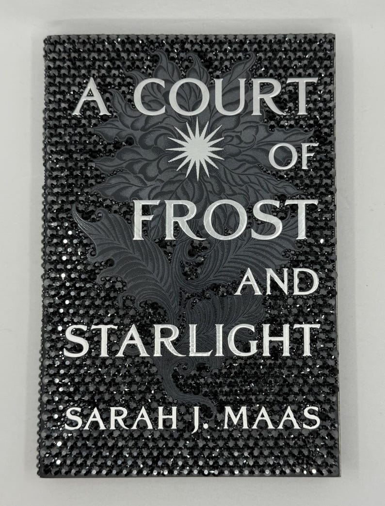 May include: A book cover for "A Court of Frost and Starlight" by Sarah J. Maas. The cover is black and encrusted with small, sparkling black gems. The title and author's name are in white, with a detailed gray floral design.