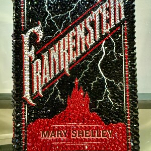 May include: A decorative piece featuring the title "Frankenstein" with a black background and white lightning bolts. The title and author's name, Mary Shelley, are in a sparkling, gem-like font. The piece has a red and black border.