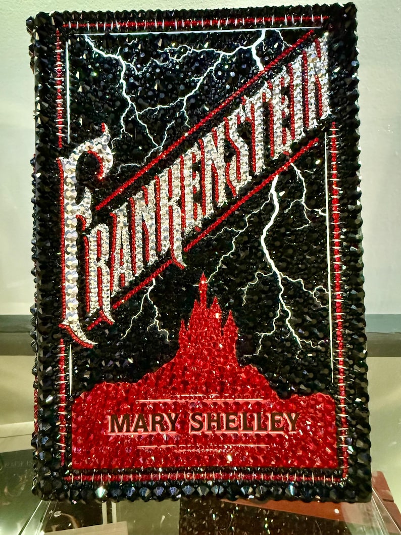 May include: A decorative piece featuring the title "Frankenstein" with a black background and white lightning bolts. The title and author's name, Mary Shelley, are in a sparkling, gem-like font. The piece has a red and black border.
