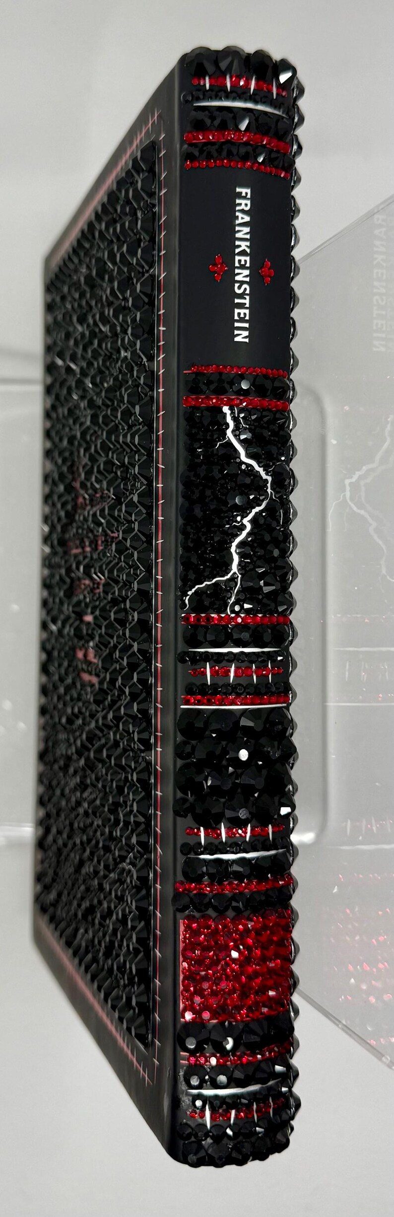 May include: Black book cover with the title "Frankenstein" in white. The spine is decorated with red and black crystals, and a white lightning bolt design. The cover is covered in black crystals.