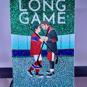 May include: A rectangular artwork with the text "THE LONG GAME" in white, featuring two hockey players facing each other. The background is a sparkling teal, with a blue and red color scheme. The artwork is likely a decorative piece.
