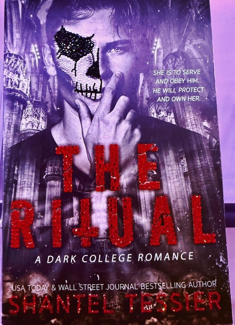 May include: Book cover for "The Ritual: A Dark College Romance" by Shantel Tessier. The cover features a man with a skull design on his face, with the title in large, red, glittery letters. The background is a dark, gothic-style image.