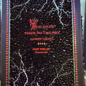 May include: A black decorative book cover with a sparkling, textured surface and white lightning bolt designs. Red text reads "You are my creator, but I am your master-obey!" and "Mary Shelley, Frankenstein."