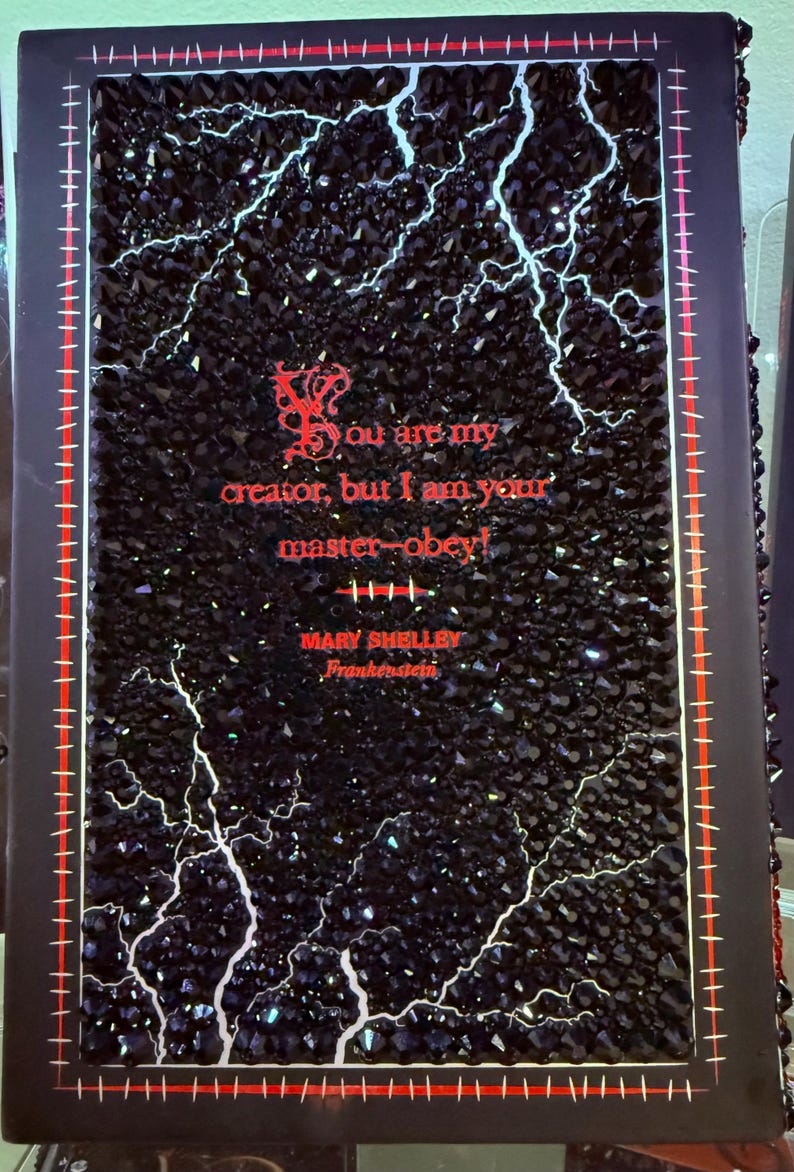 May include: A black decorative book cover with a sparkling, textured surface and white lightning bolt designs. Red text reads "You are my creator, but I am your master-obey!" and "Mary Shelley, Frankenstein."