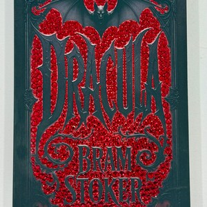 May include: A dark teal book cover with the title "DRACULA" in large, stylized letters, filled with red gems. The author's name, "BRAM STOKER," is below. A bat graphic is at the top, and the cover has an ornate border.