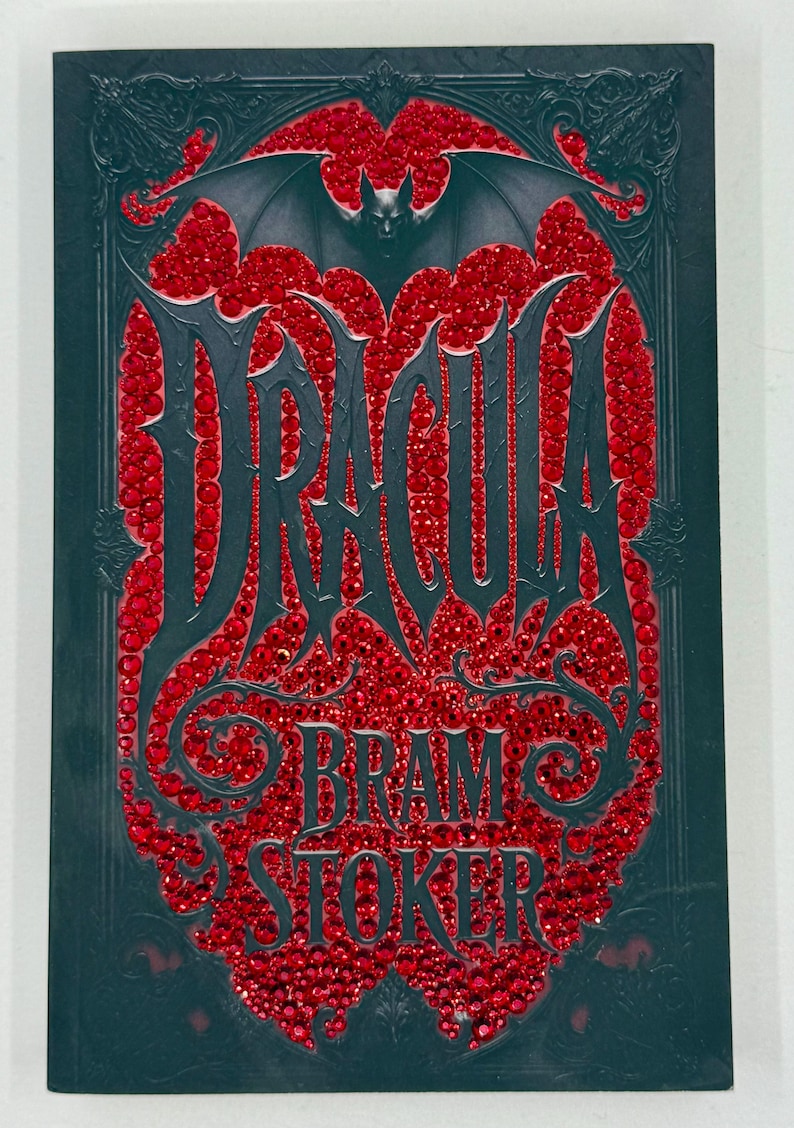 May include: A dark teal book cover with the title "DRACULA" in large, stylized letters, filled with red gems. The author's name, "BRAM STOKER," is below. A bat graphic is at the top, and the cover has an ornate border.