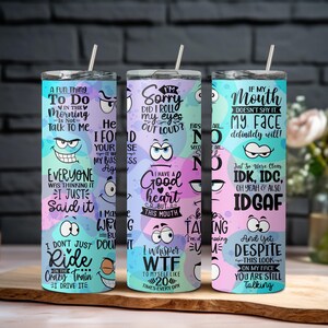 May include: Three stainless steel tumblers with humorous phrases and cartoon faces. The tumblers have a straw and are decorated with pastel colors and various quotes. The background is a wooden cutting board.