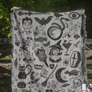 May include: A gray woven throw blanket with a black and white pattern of various spooky and witchy imagery, including a cat, a snake, a skull, a witch, a coffin, a crescent moon, a broom, a potion bottle, a spider, a bat, a hand, a dagger, a feather, a candle, a book, a heart, a pumpkin, a crystal, a mirror, a dice, a feather, and a snake. The blanket has fringe on all sides. The text "THE OWLS GLOW" is visible in the bottom right corner of the image.