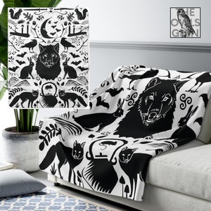 May include: A black and white throw blanket featuring a repeating pattern of a wolf, foxes, rabbits, bats, and other forest elements. The blanket is draped over a sofa, showcasing the detailed design. The Owls' Glen logo is visible in the background.